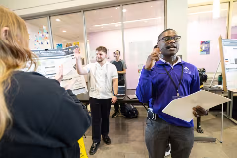 A student waves to the audience after presenting his poster at Showcase Day while a man with a UAlbany Student Affairs shirt speaks into a headset microphone.
