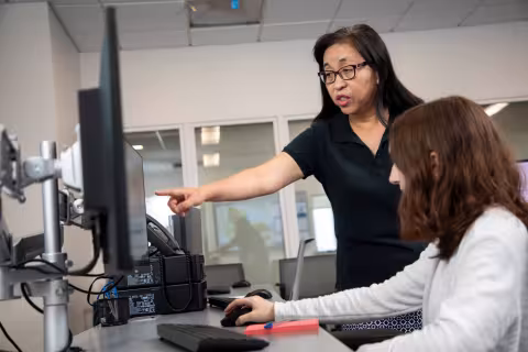 An instructor looks over a student's shoulder and points to their computer screen.