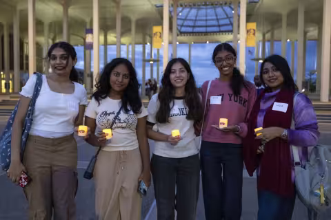 Five young woman, all with long dark hair, smile for a group photo. All are holding small shite tealight candles bearing the UAlbany logo. 