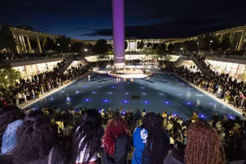 Hundreds of students gathered around UAlbany’s main fountain. The sky is dark and blue LED lights shine up from the bottom of the fountain’s reflecting pool. The base of the carillon is lit purple. 