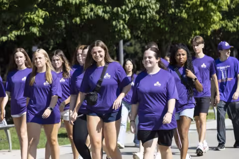 A group of students wearing purple UAlbany T-shirts walk to convocation together. A young woman in the front, middle is smiling at the camera. 
