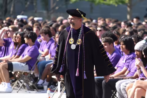 UAlbany’s President Havidán Rodríguez, smiling, wearing a black robe and black tasseled cap, approaches the podium deliver remarks at Convocation.