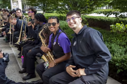 Member of UAlbany’s marching band sit in a line waiting to play at convocation. Two members on the end, one holding a saxophone, smile at the camera. 