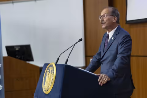 A man in a blue suit and glasses speaks at a lectern.