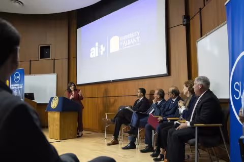 A woman in a red suit stands at a lectern and gestures to the screen behind her.