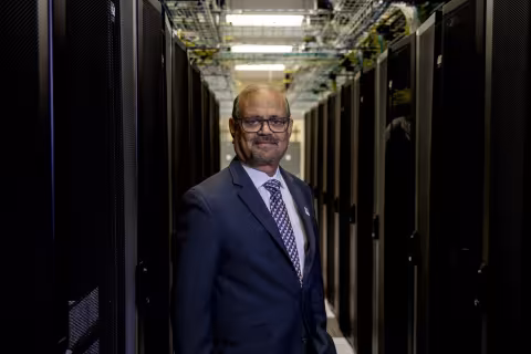 A man in a dark suit facing the camera between two long rows of computers.