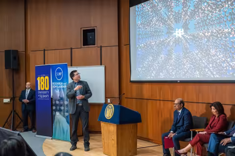 A man in a suit points to the projection screen behind him featuring a complex molecular pattern.