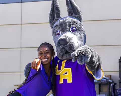A student wearing a purple shirt smiles and poses with UAlbany mascot Damien the Great Dane as they both point at the camera.