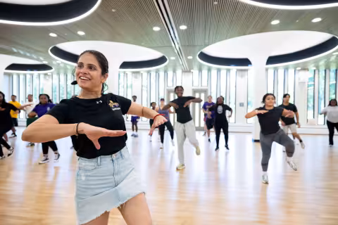 A dance instructor smiles and gestures while leading a class inside a group exercise room with a wood floor, a mirrored wall and bright white columns.