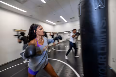 A student wearing boxing gloves moves to punch a boxing bag inside a group exercise room with boxing and kickboxing equipment.