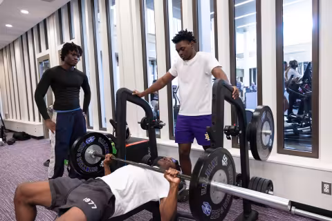 A student does bench presses while a second students spots him and a third waits for his own turn.