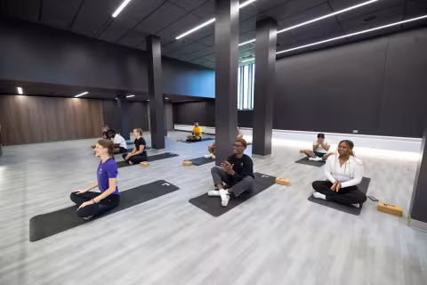 An instructor leads a yoga class inside a group exercise room with a wood floor, a mirrored wall, a dance bar and gray columns.