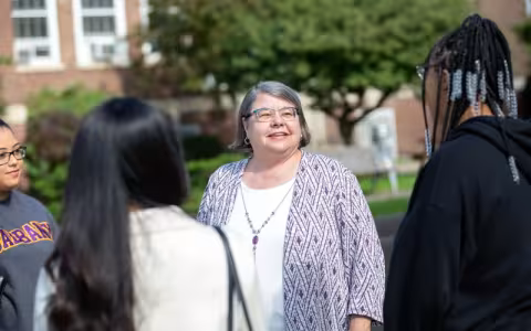 Four people stand together outdoors. A woman in the center smiles at three female students who are facing her. The woman has short gray hair and is wearing a purple and white patterned jacket and glasses. A tree and red brick building are in the background.
