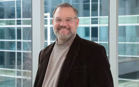 Portrait of Scott Tenenbaum: A man wearing clear-rimmed glasses, a dark brown blazer and tan turtleneck sweater stands, smiling, in front of a wall of glass windows.