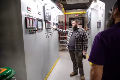 A man stands gesturing to a massive control panel with numerous switches and digital screens.