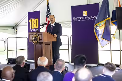 A man stands at a wooden lectern on a stage surrounded by purple banners.