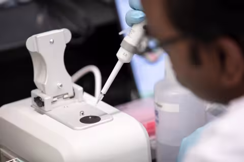 A man wearing glasses and gloves dispenses a substance from a pipette onto a small dark platform on the top of a white plastic lab instrument.