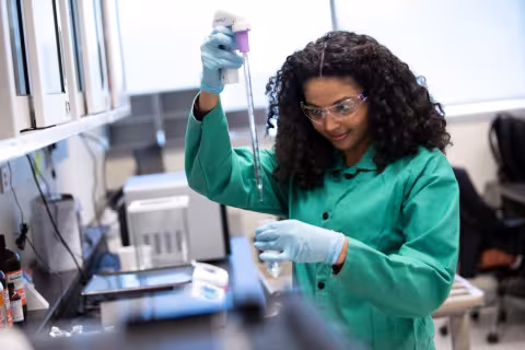 A young woman with dark curly hair wearing protective glasses and a green lab coat uses a pipette to transfer a clear liquid into a clear plastic test tube. 