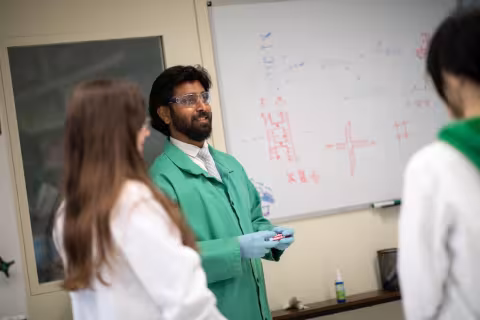 A man with dark hair wearing protective glasses, a green lab coat and gloves stands in front of a whiteboard addressing two students wearing white lab coats. 