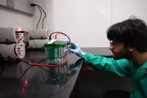 A young man wearing protective glasses and a green lab coat adjusts a gel electrophoresis device comprised of a clear plastic box with a green lid, to which two electric cords (one black, one red) are attached. 