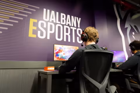 A student competes in the SUNY Esports Tournament in front of a UAlbany Esports graphic.