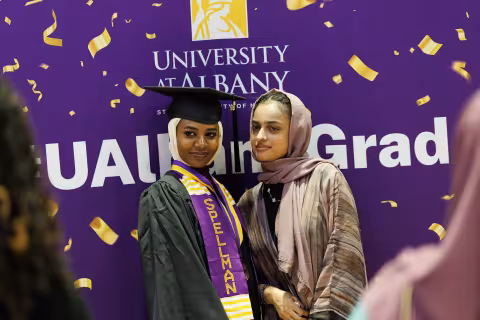Two young women, one in a graduation gown and the other in a traditional hijab, smile as their photo is taken while standing in front of a sign that says "UAlbnay Grad"