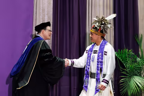 A young man in traditional Iroquois regalia - a white robe with lettering and an Iroquois head dress - shakes hands with an older man while crossing a graduation stage.