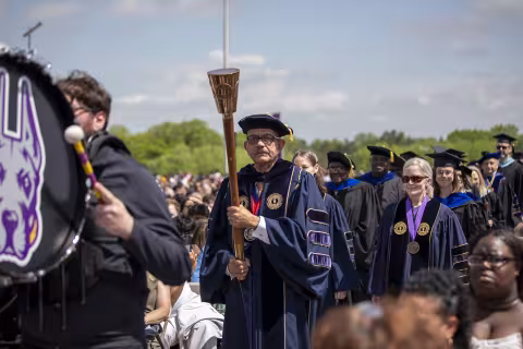 A man in fancy purple commencement robes walks with a ceremonial mace leading a parade of people dressed in similar robes outdoors against a blue sky.