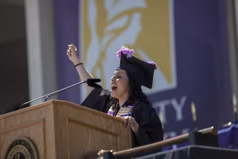 A latina woman in commencement robes speaks at a podium with a golden graphic of Minerva against a purple background behind her outdoors.