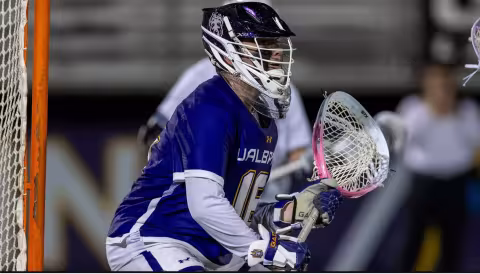 A man wearing a purple jersey and a large helmet holds a goalie lacrosse stick as he looks off in the distance.