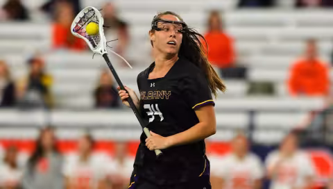 A game photo of a girl with a lacrosse stick, goggles and a yellow lacrosse ball wearing a black shirt that says UALBANY and has the number 34 on it.