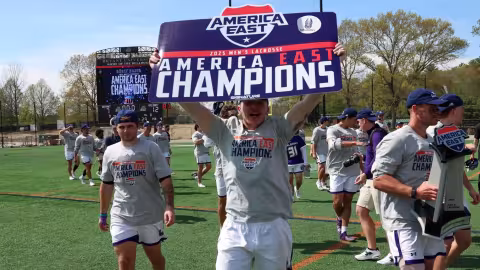 A man in a gray shirt and white shorts holds a sign that says America East Champions as his teammates walk behind him on a green athletic field during the day.