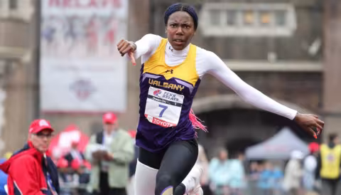 A girl wearing a white long sleeve shirt and a yellow and purple jersey jumps down the runway on an athletic track.