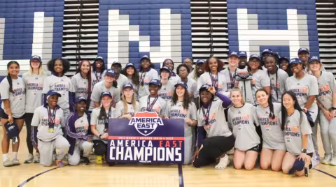 A large group of women wearing gray shirts in a gymnasium hold a sign that says America East Champions.