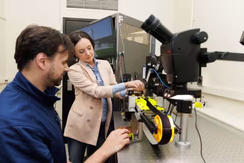 A man wearing a blue shirt and a women wearing a peach blazer tinker with a piece of lab equipment that features a microscope and a small black and yellow conveyor belt powered by a hand crank. 