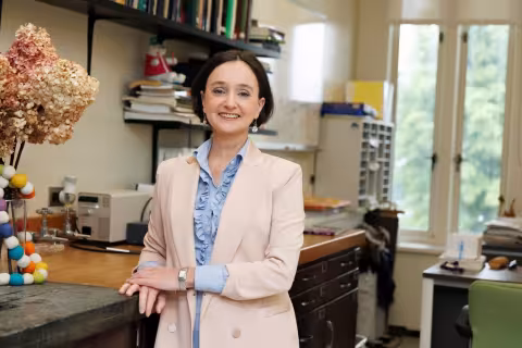 A woman with short brown hair wearing a peach blazer stands in an office, smiling at the camera. Bookshelves, dried flowers and a window can be seen behind her.  