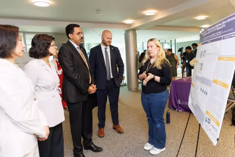 A student explains her research poster to three people dressed in suits. They are standing in an open space with other people milling in the background.