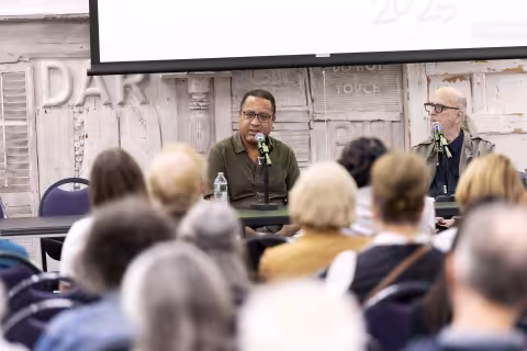 A Black man in the backgroudn talks to a crowd of people in the foreground indoors in a discussion setting.