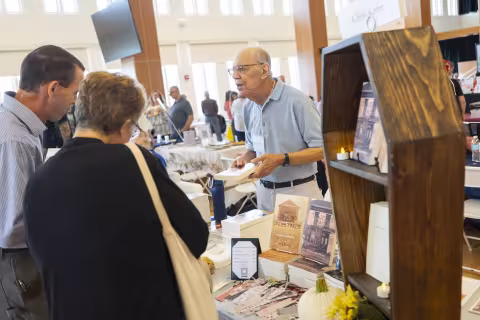 A older man leans over a table of books and talks with an older couple indoors.