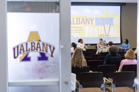 a logo with UALBANY written on it is in foreground as people in the background talk at a table with the words Albany Book Festival behind them on a screen indoors.
