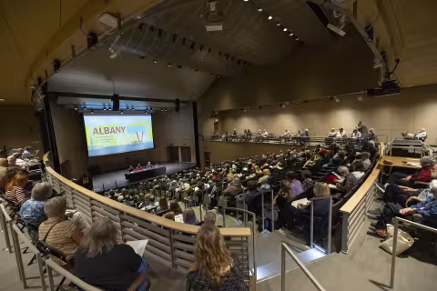 A large audience of people in an auditorium listen to three people seated on a stage in front of a sign that reads Albany Book Festival