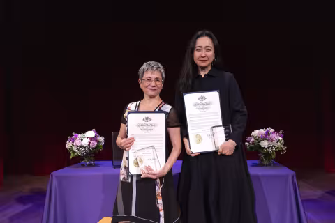 A short woman in a colorful blouse and a tall woman in a dark dress stand with proclamations of State Poet and State Author against a black backdrop and a table with a purple cover.