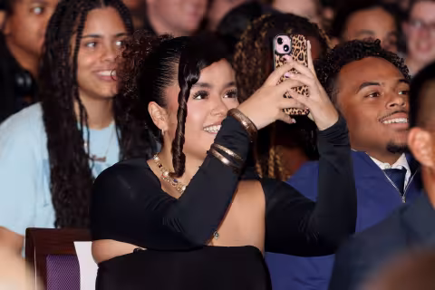A young Latina woman with dark hair and dark eyes in a black dress is seated among a crowd indoors and takes a picture with her mobile phone of something off screen in front of her.