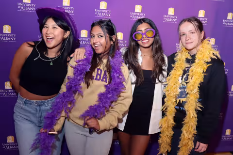 A group of four young women wear purple and gold festive costumes while having their photo taken in front of a stand that has the words "University at Albany" in small writing behind them.