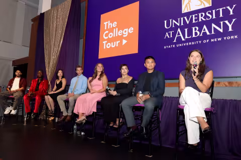 A group of students in colorful outfits sit on high chairs holding microphones in a line as part of a panel discussion, as a display behind them reads "The College Tour" and "University at Albany".