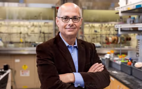 A portrait of a man in a dark jacket standing with his arms crossed inside a chemistry lab. 