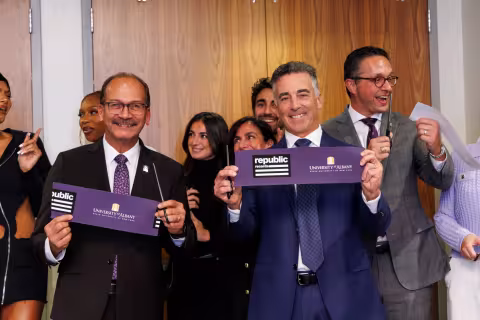 UAlbany President Havidan Rodriguez holds a ribbon next to Avery Lipman, '88, indoors along a wall with wood paneling.