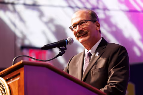 A man in a gray suit and purple tie with glasses and a mustache speaks from a lectern indoors in an auditorium.