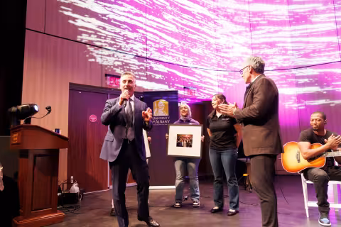 A group of people stand on a stage as purple lights appear on the screen and two individuals are given a sign in their honor.