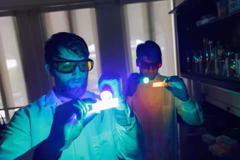 Two men in lab coats use handheld lights in a darkened lab to illuminate specimen slides that they are holding.